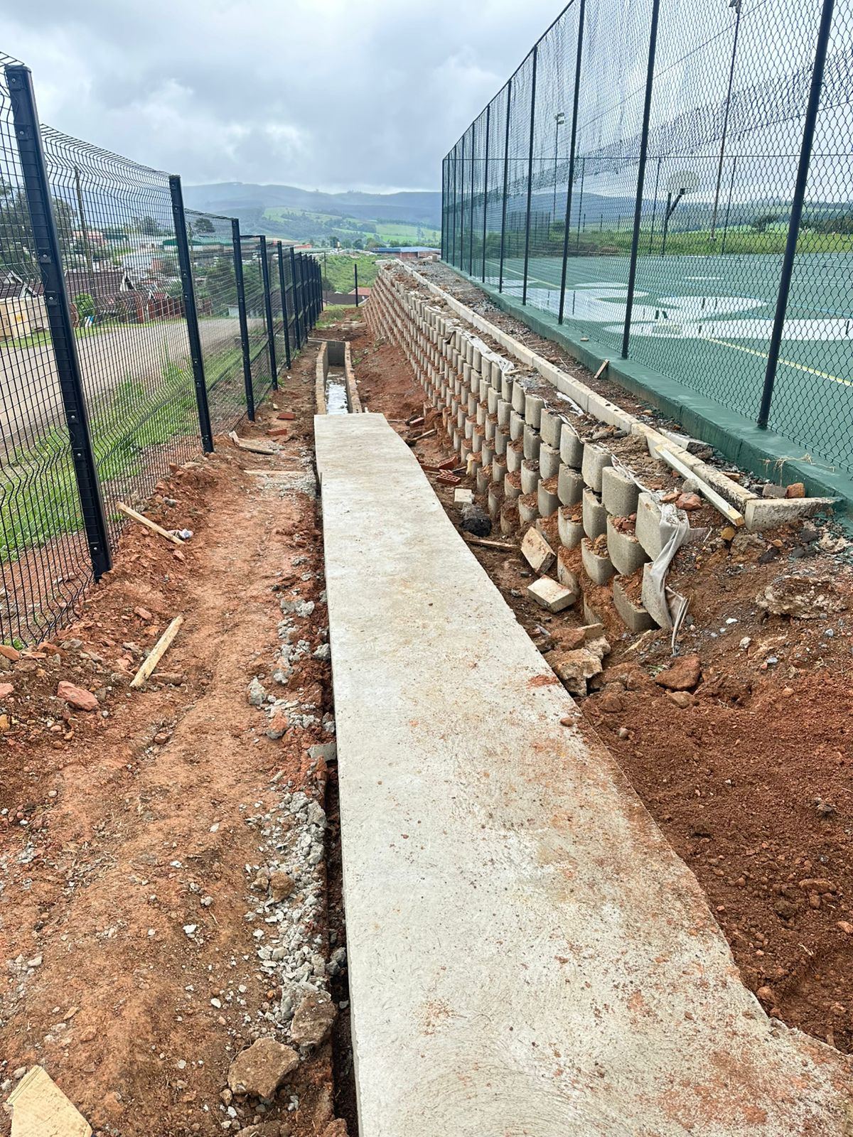 Mesh fencing, retaining wall and concrete drainage channel at a sports court